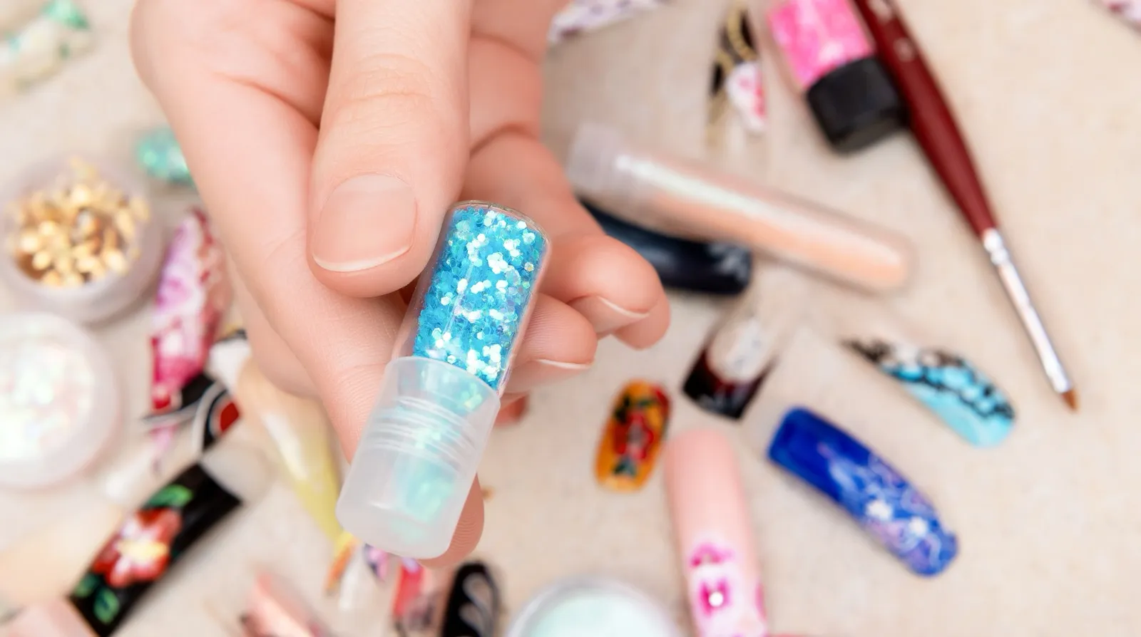 A hand holding a small vial of blue glitter over a table of finished nail art tips, brushes and pots of pigment
