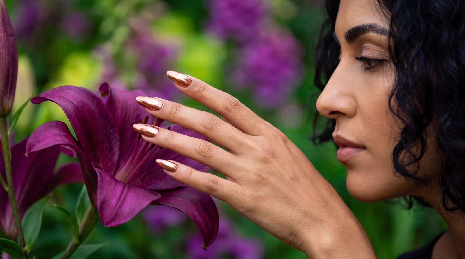 A woman in profile touching a deep purple lily, her almond nails finished in a rose gold chrome mirror