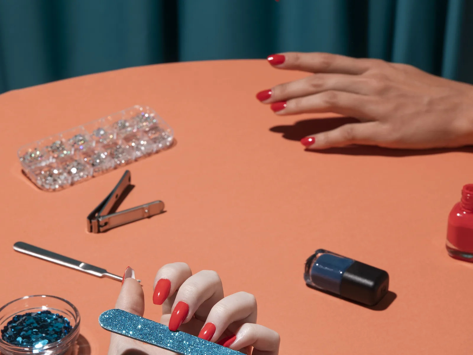 Close-up of a manicure in progress: red gel nails being filed with a glitter file, a red polish bottle open beside them on an orange table