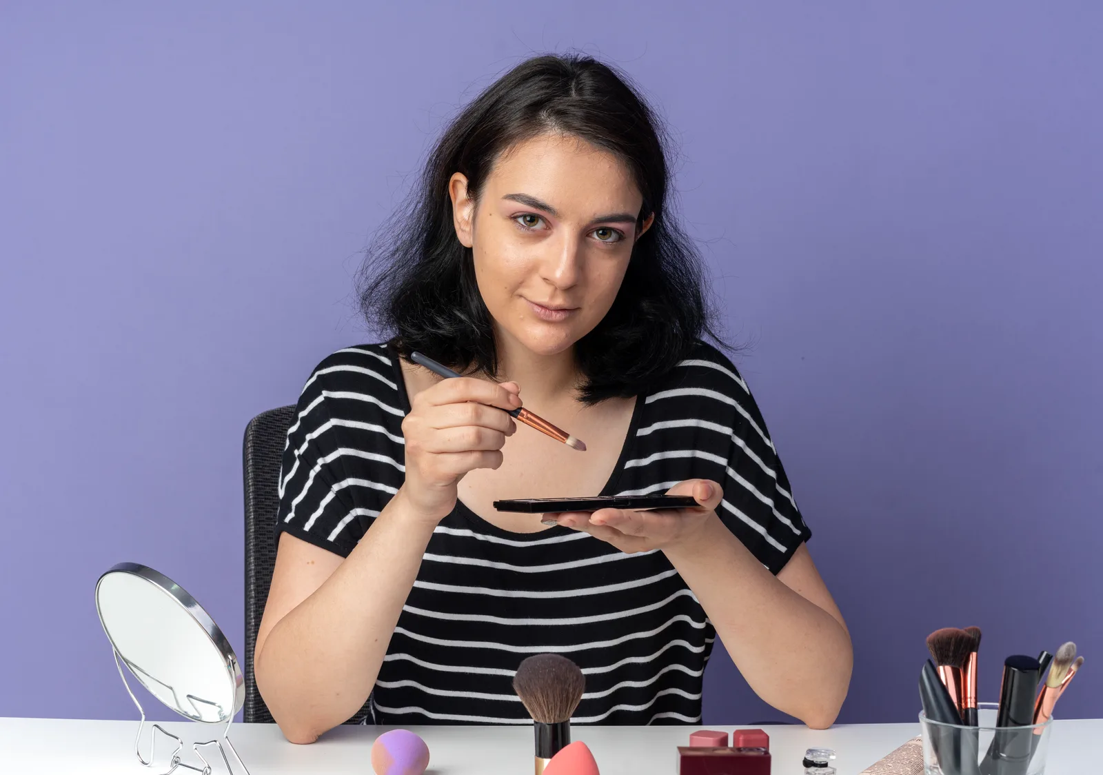 A young woman at a table holding a fine brush over a palette, with a mirror and a pot of brushes beside her against a blue wall
