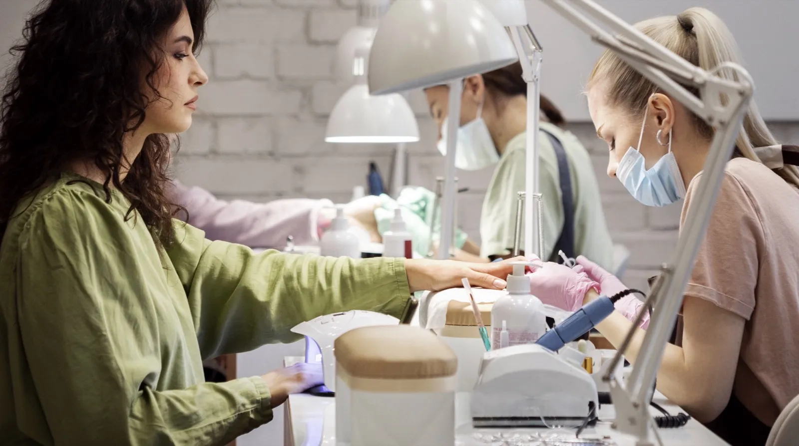 A client rests a hand under a curing lamp at a salon desk while a masked technician in pink gloves works on a hand with a handheld nail drill