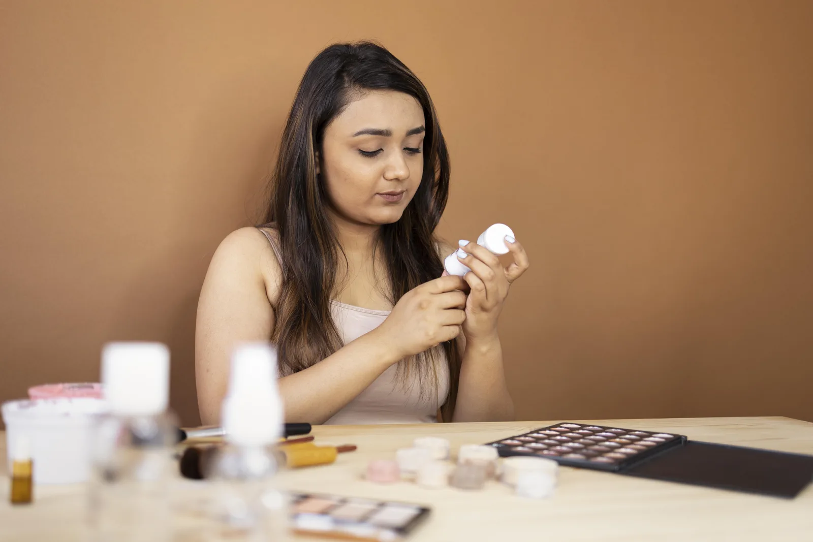 A woman at a desk reading the label on a small white pot, with palettes, pots and a spray bottle laid out in front of her