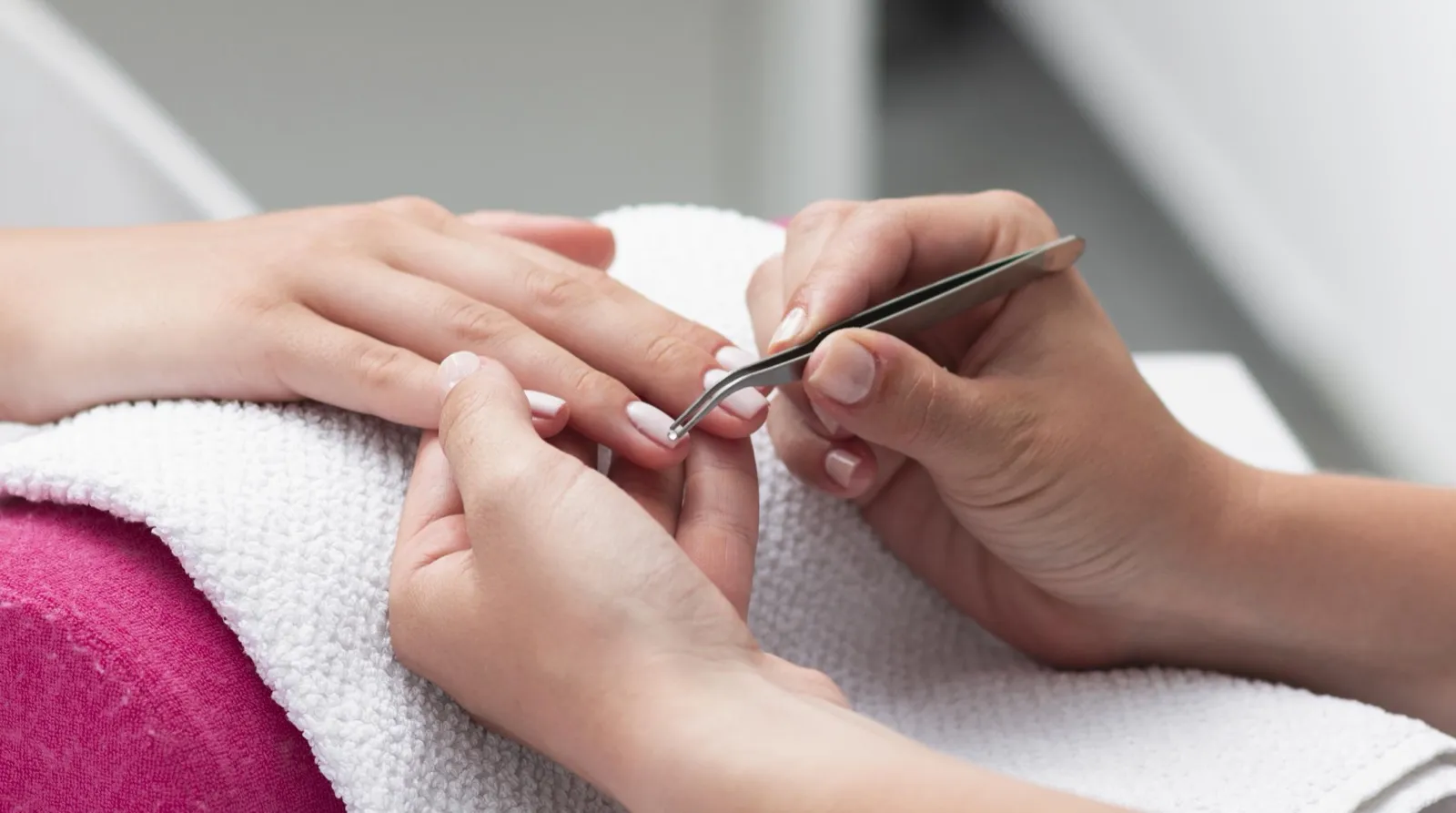 A technician using tweezers to press a pale nail tip onto a client's fingertip while the hand rests on a white towel